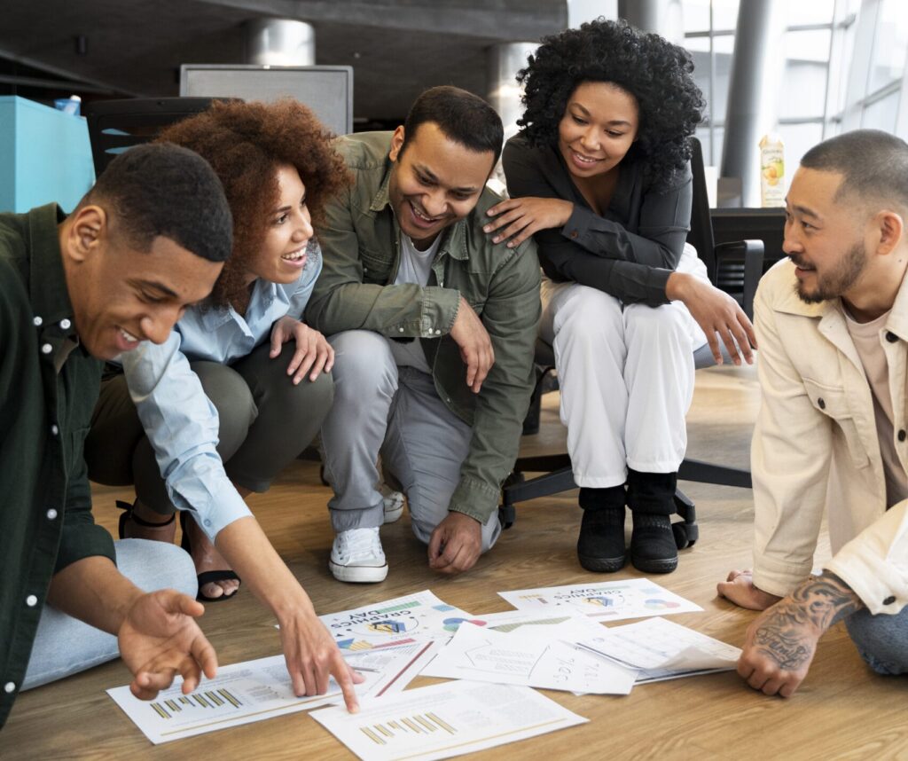 Grupo diverso de jóvenes profesionales reunidos en una oficina moderna, colaborando sobre documentos y gráficos en el suelo.